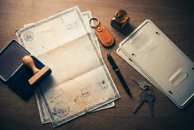 Title documents, a rubber stamp, keys, and a blank license plate arranged on a desk
