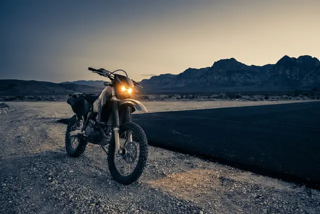 Dirt bike with its headlight lit where a gravel trail meets new pavement at dusk
