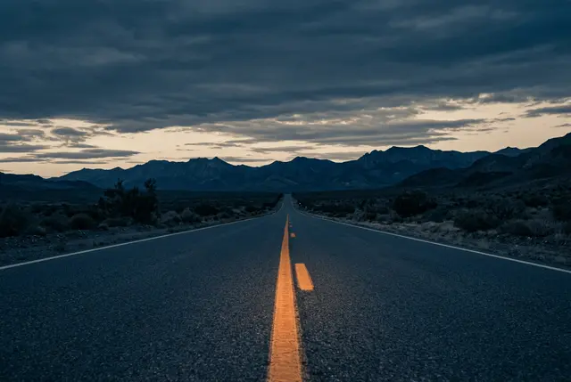 Empty two-lane highway with a glowing amber centerline heading toward mountains at dusk